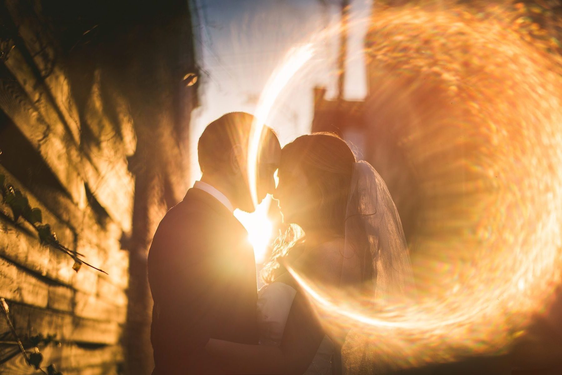 A bride and groom leaning in to kiss in silhouette, ringed by a circle of golden sun flare against a stone wall