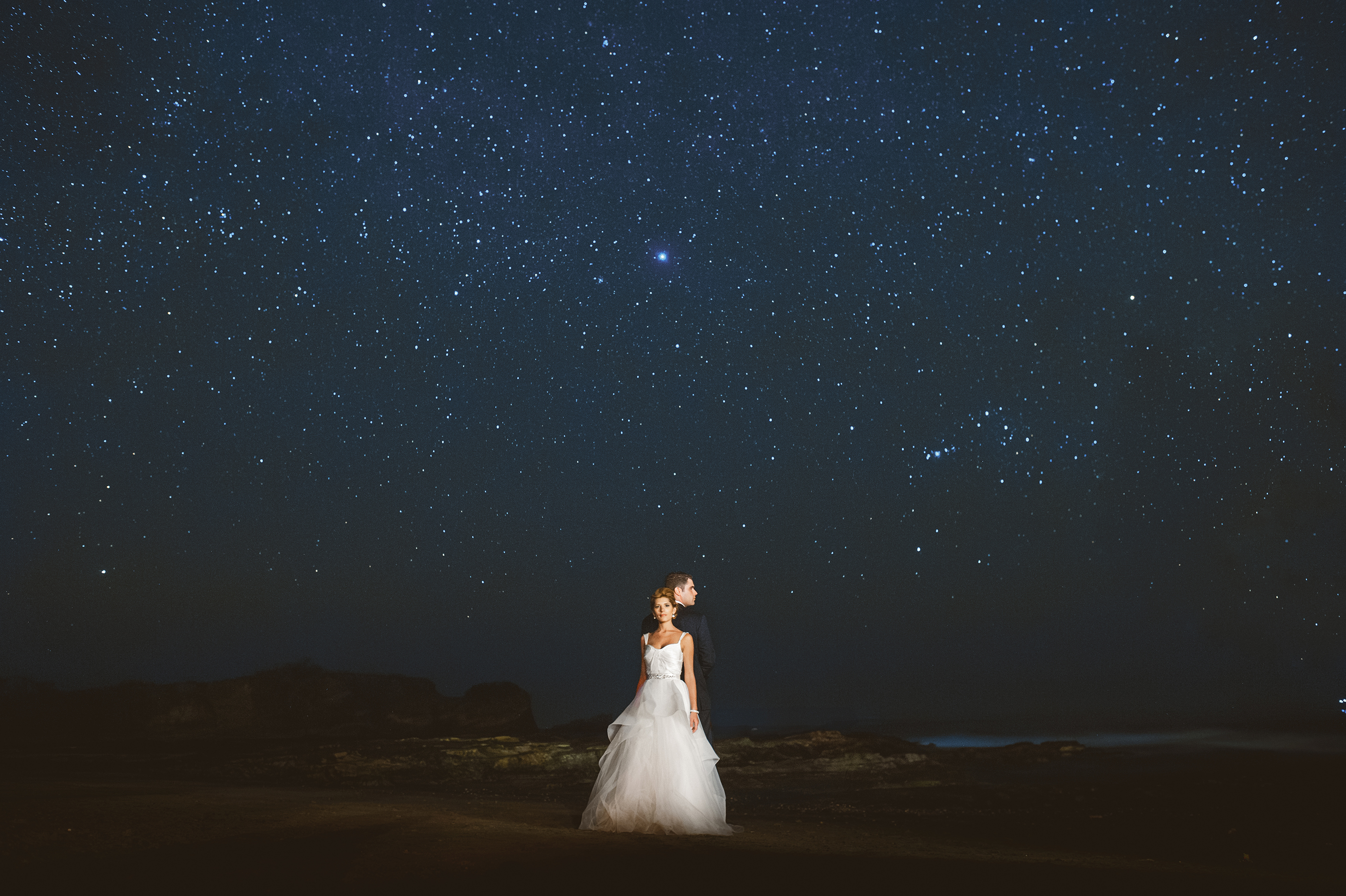 A bride and groom lit against a night sky full of stars on open rocky ground